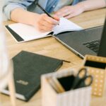 Adult writing notes on a notebook while working on a laptop in a cozy home office environment.